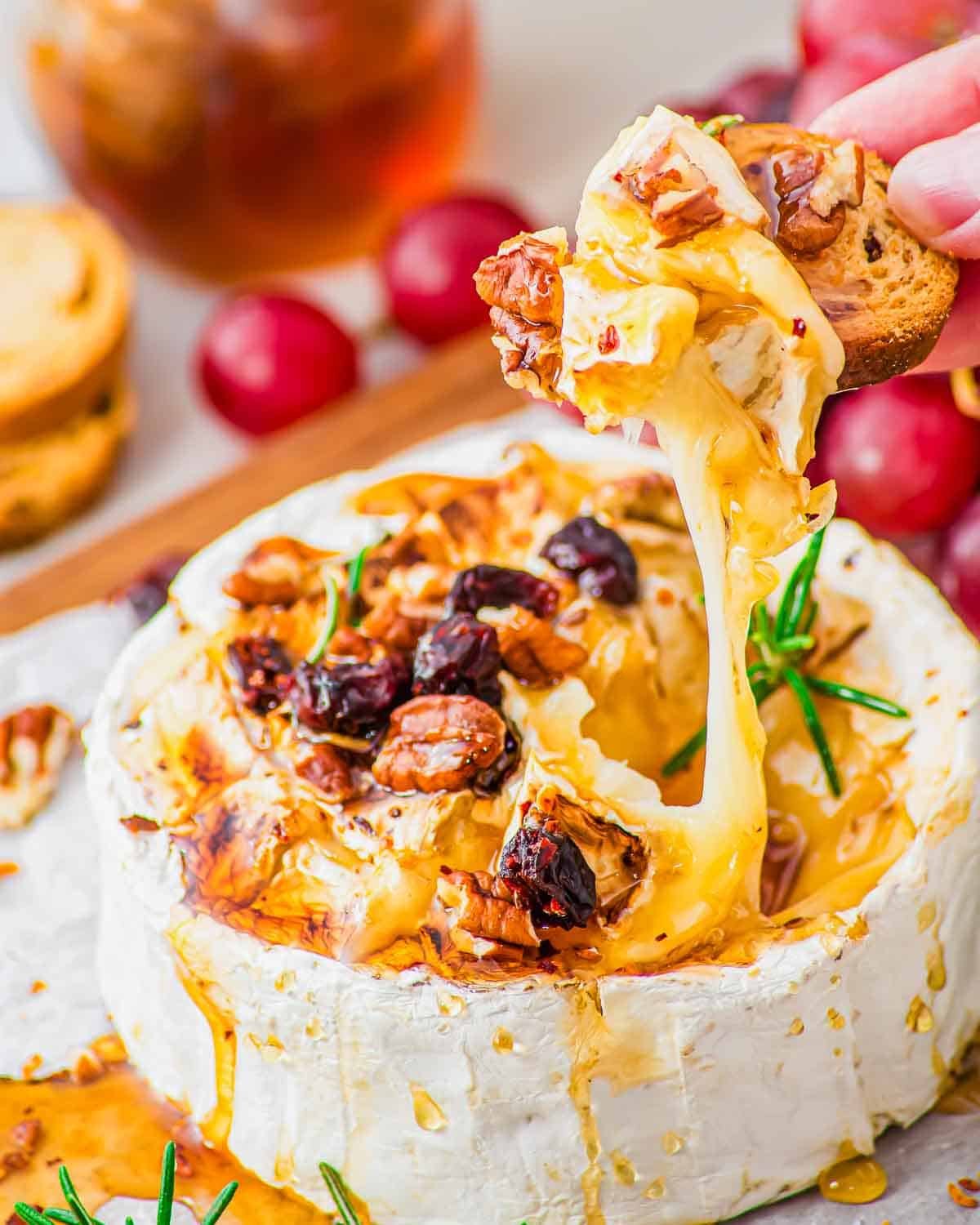 A hand dips a toasted baguette slice into warm, gooey baked brie topped with honey, nuts, and dried cranberries. Fresh rosemary and grapes are seen nearby on the serving board.