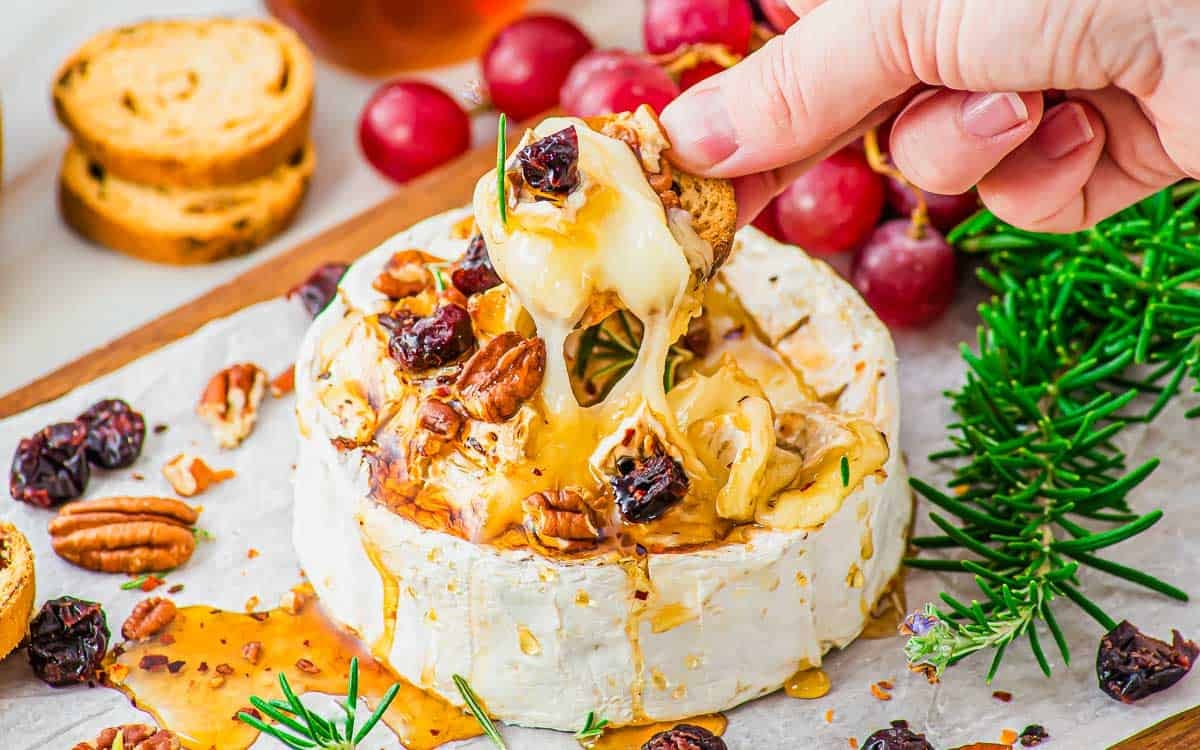 A hand dips a cracker into melted baked brie topped with pecans, dried cranberries, honey, and rosemary. Grapes, more toasts, and fresh rosemary are on the side.