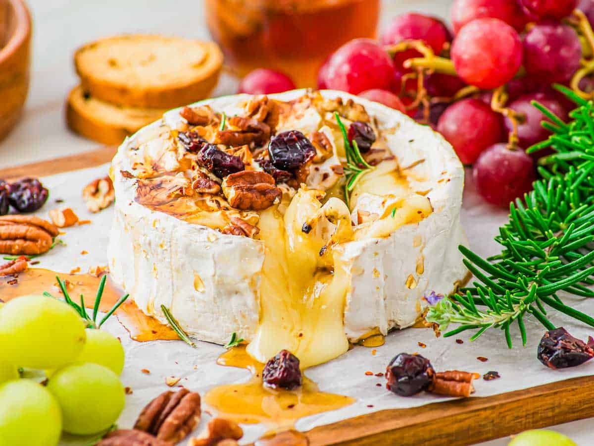 A wheel of baked brie cheese with honey is topped with pecans, dried cranberries and fresh rosemary, with cheese melting out. Fresh grapes, crackers, and rosemary sprigs are arranged nearby.