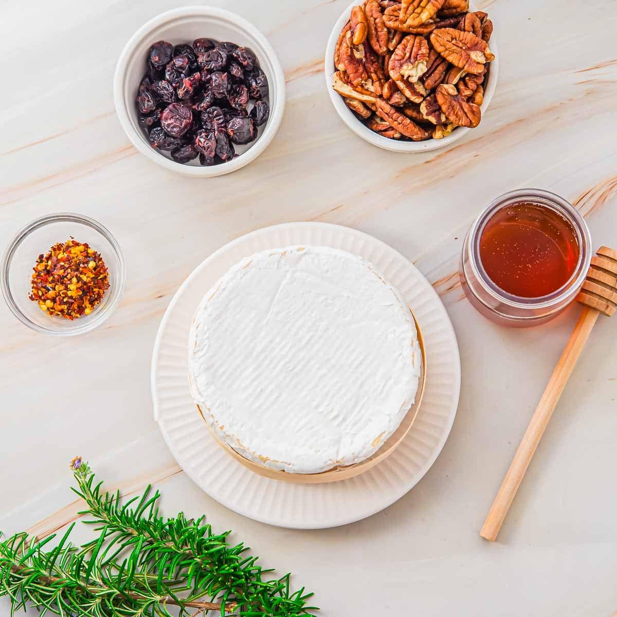 A round wheel of brie cheese on a white plate, surrounded by bowls of dried cranberries, pecans, crushed red pepper, a jar of honey with a dipper, and a sprig of fresh rosemary on a light surface.