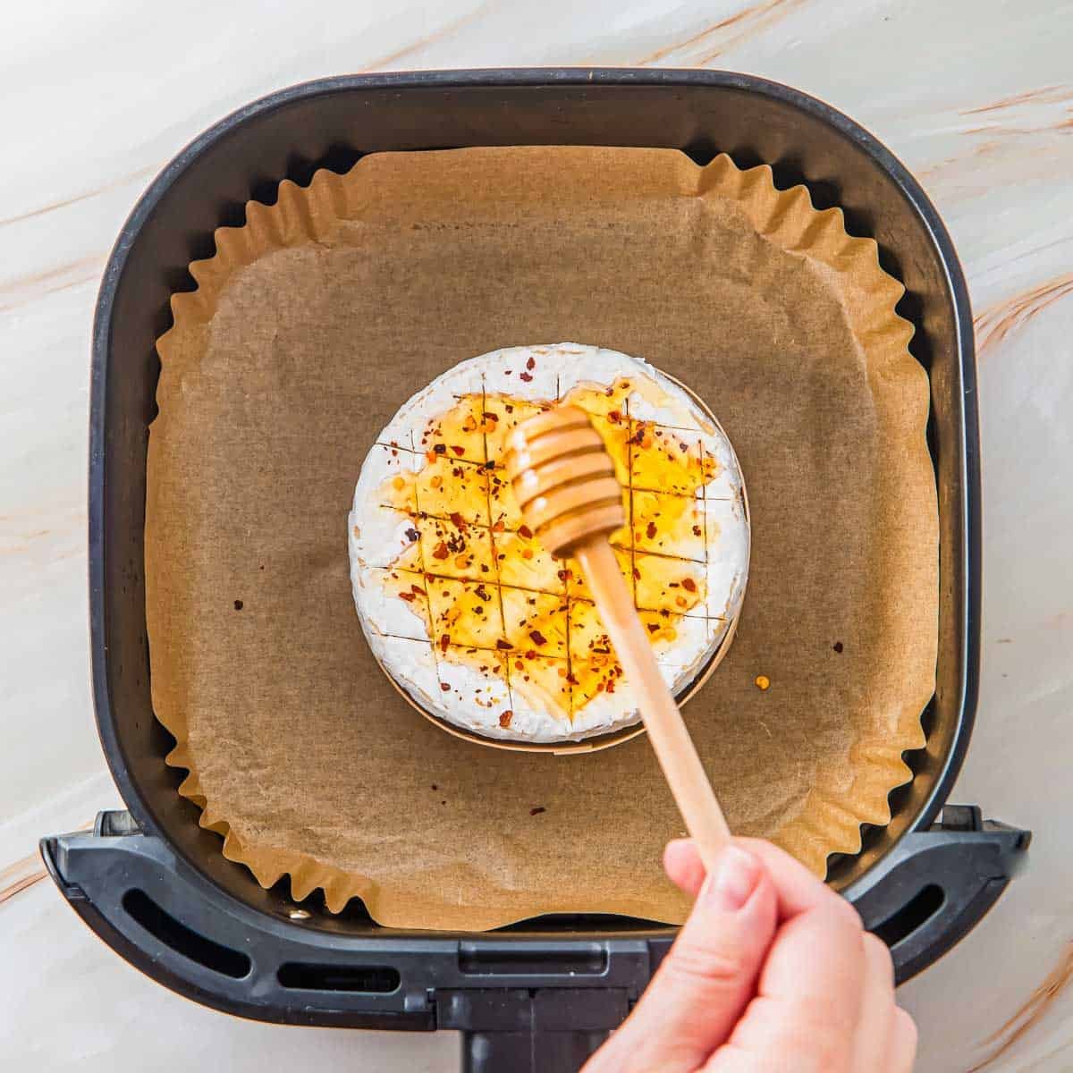 A hand holds a honey dipper over a round wheel of brie cheese scored with a crosshatch pattern and sprinkled with chili flakes, placed on parchment paper inside an air fryer basket.
