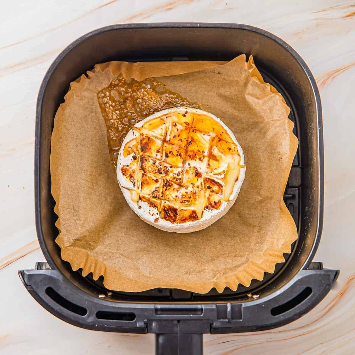 A round wheel of baked brie cheese with a scored top sits on parchment paper inside an air fryer basket, melted cheese bubbling out on one side.