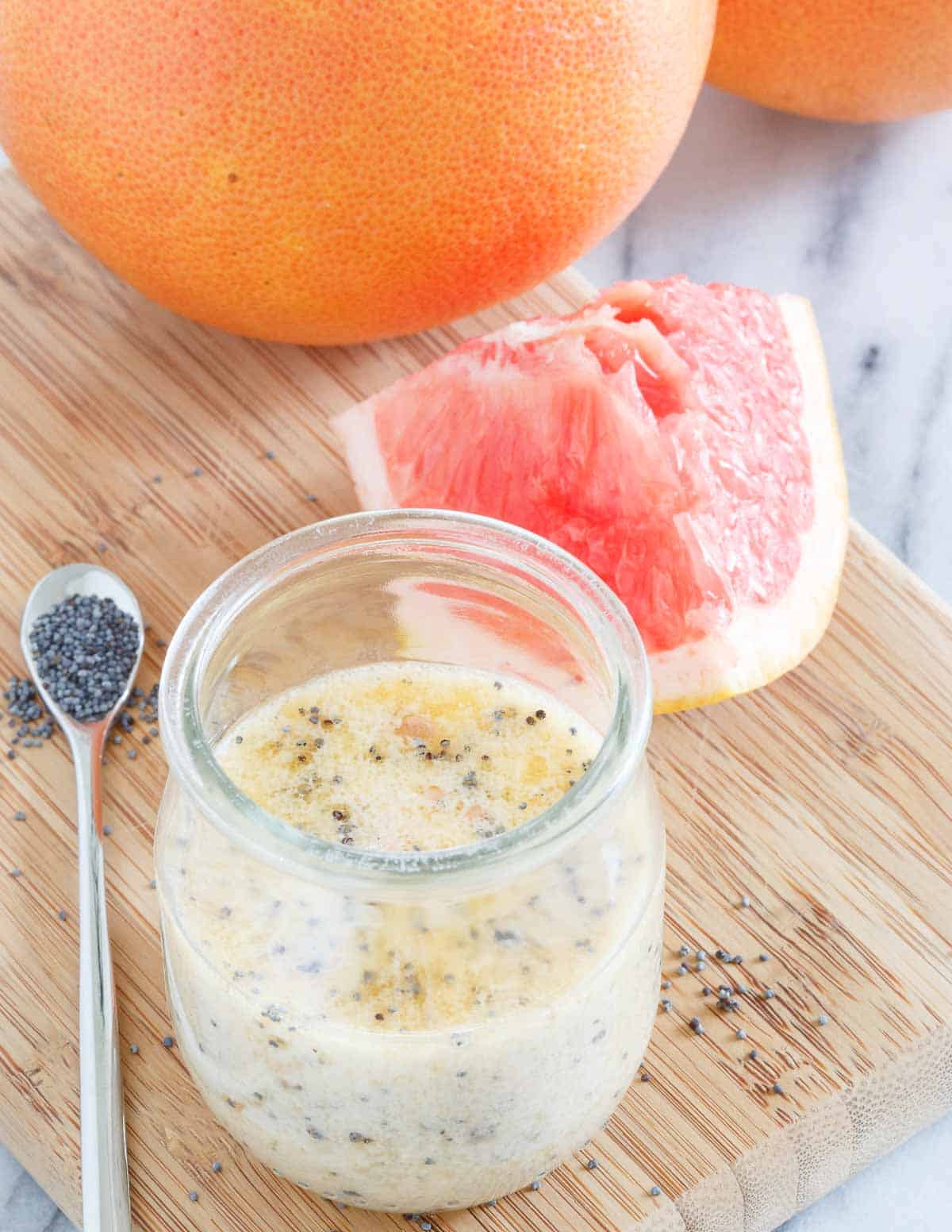 A small glass jar with creamy citrus dressing containing poppy seeds sits on a wooden board next to a spoon of poppy seeds and a wedge of pink grapefruit. Whole grapefruits are in the background.