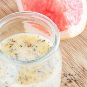 A glass jar filled with grapefruit poppyseed dressing sits on a wooden surface next to a fresh slice of pink grapefruit. Some poppyseeds are scattered on the table.