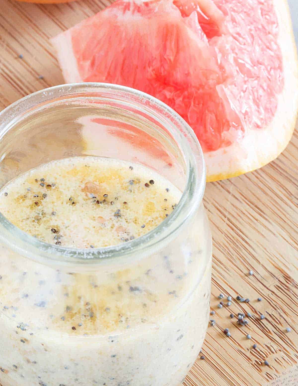 A glass jar filled with grapefruit poppyseed dressing sits on a wooden surface next to a fresh slice of pink grapefruit. Some poppyseeds are scattered on the table.