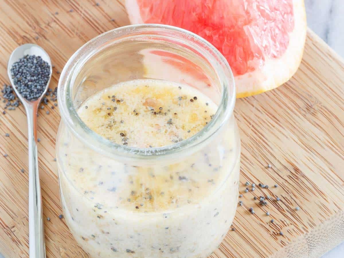 A glass jar of creamy citrus poppy seed dressing sits on a wooden board next to a sliced grapefruit and a spoonful of poppy seeds, with some seeds scattered on the board.