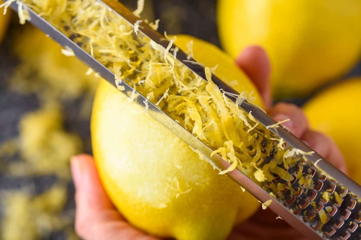 A close-up of a hand using a metal grater to zest a lemon, with fresh lemon zest collecting on the grater and whole lemons blurred in the background.