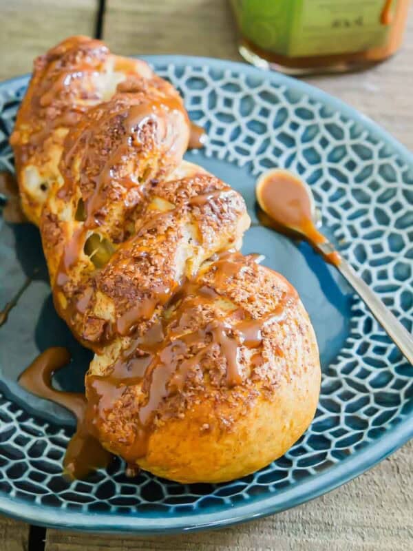 A pretzel twist is drizzled with caramel sauce sitting on a blue patterned plate, with a spoon and a jar of sauce in the background.