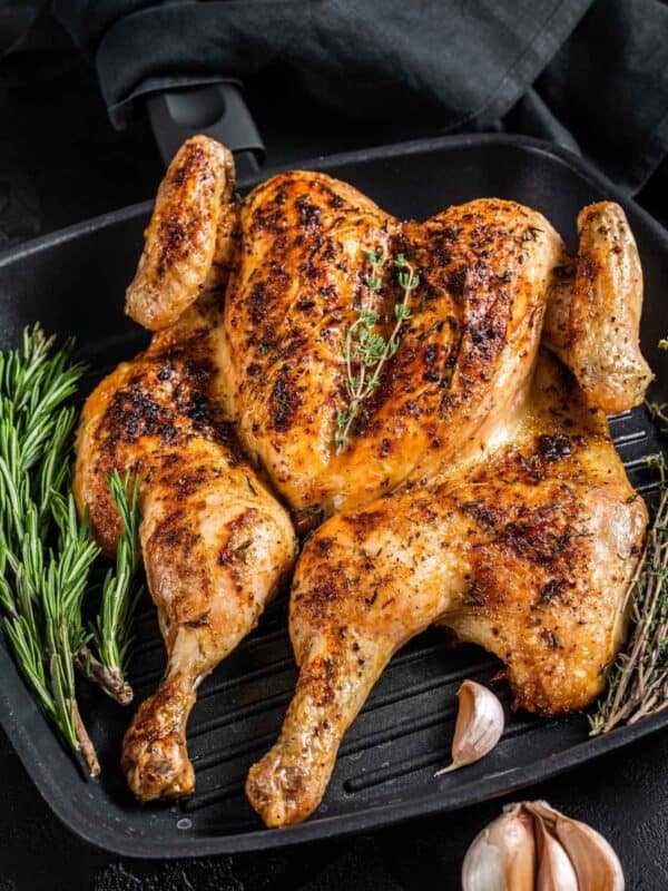 A whole roasted spatchcock chicken with crispy, golden-brown skin sits on a grill pan, garnished with fresh rosemary, thyme, and whole garlic cloves. A black cloth is draped in the background, highlighting the succulent spatchcock chicken.