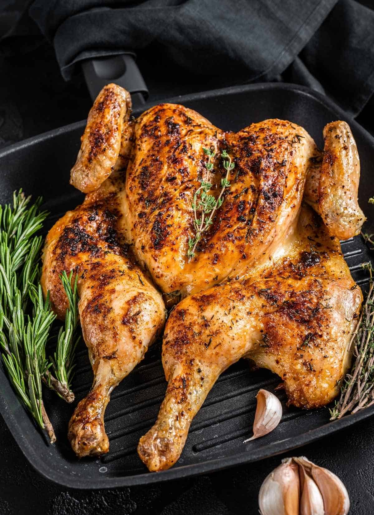 A whole roasted spatchcock chicken with crispy, golden-brown skin sits on a grill pan, garnished with fresh rosemary, thyme, and whole garlic cloves. A black cloth is draped in the background, highlighting the succulent spatchcock chicken.