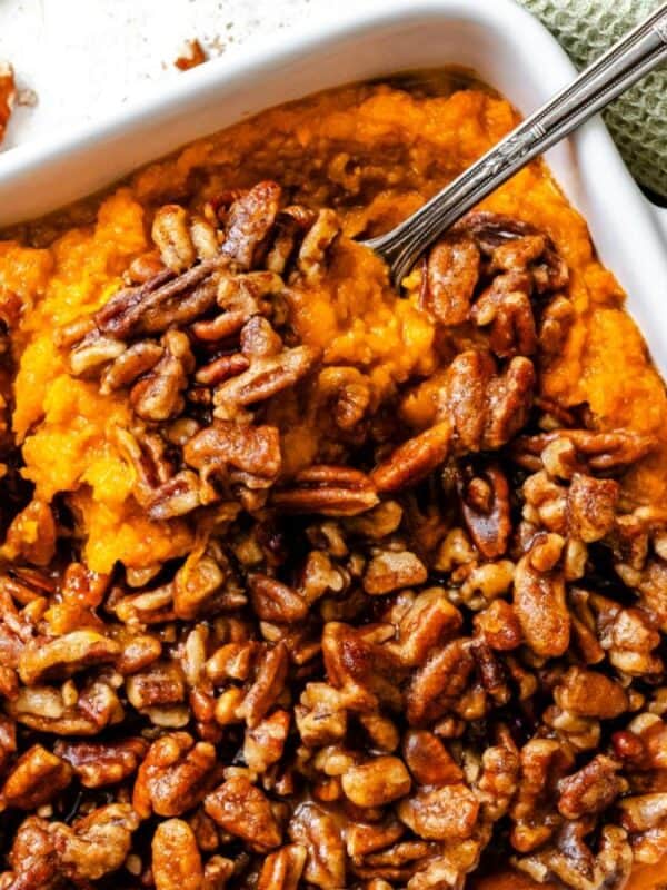 A white baking dish filled with sweet potato casserole topped with glazed pecans, with a serving spoon inside. A bowl of pecans and a green towel are nearby on the table.