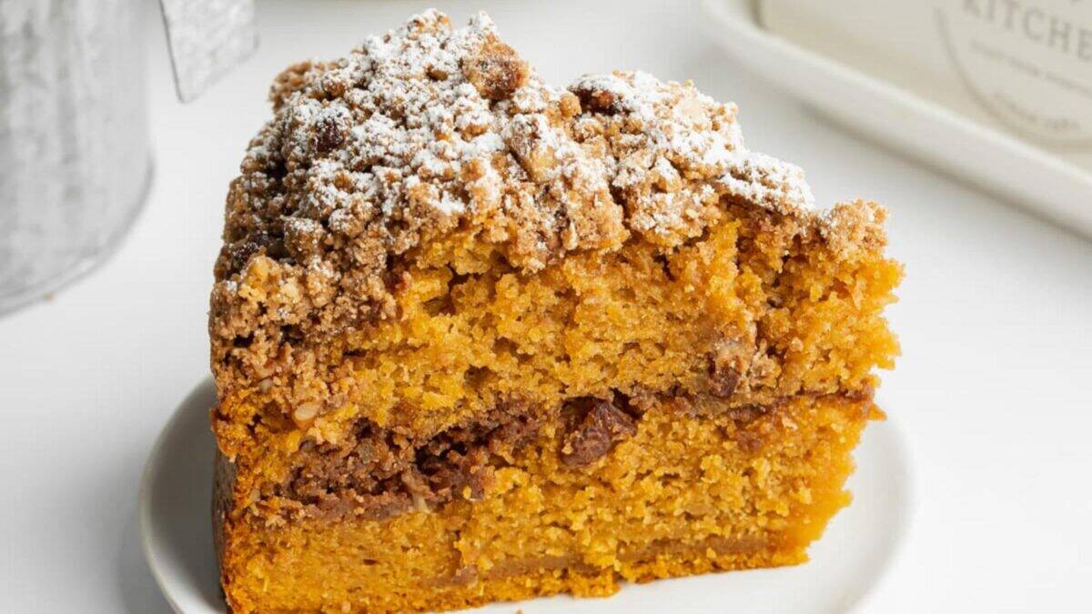 A close-up of a slice of moist pumpkin coffee cake with a crumbly streusel topping and powdered sugar, served on a white plate against a light background.