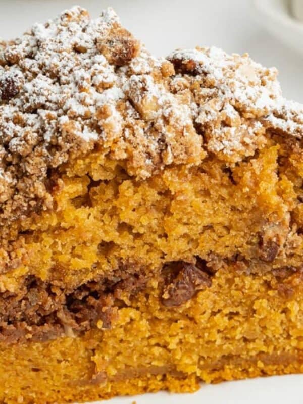 A close-up of a slice of moist pumpkin coffee cake with a crumbly streusel topping and powdered sugar, served on a white plate against a light background.