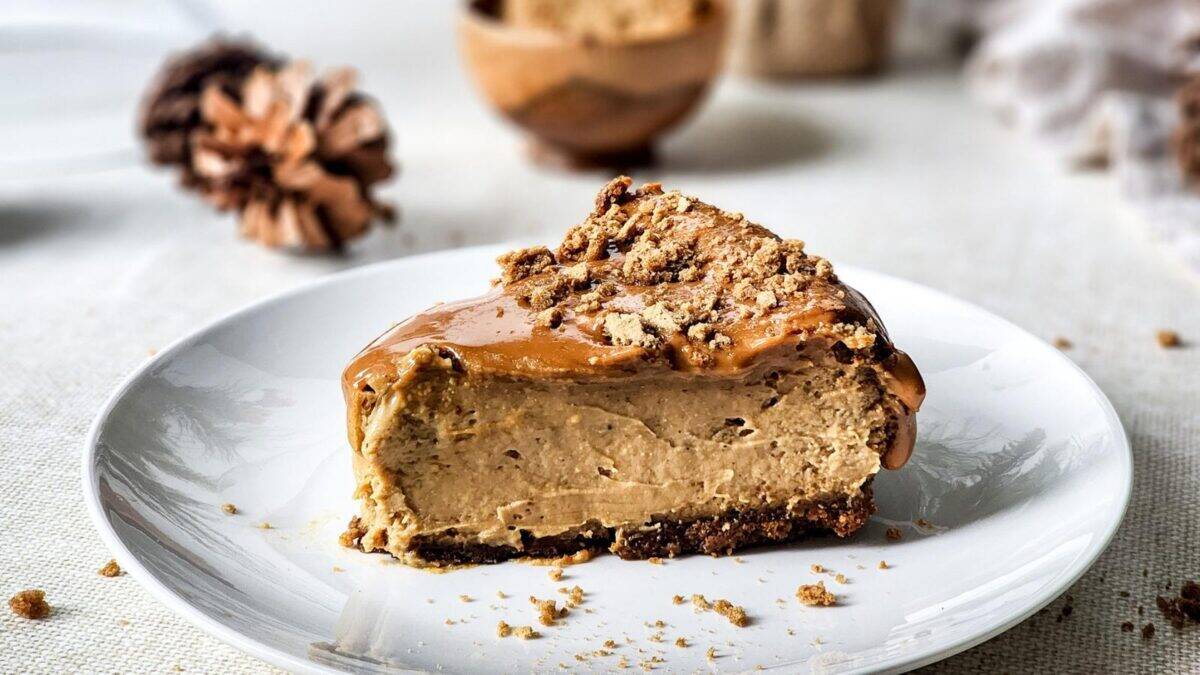 A slice of creamy cheesecake with a crumbly crust and caramel topping sits on a white plate, with crumbs scattered around and a pinecone decor in the blurred background.