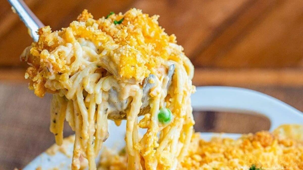 A close-up of a cheesy baked casserole being lifted with a spoon, showing creamy noodles, green peas, and a golden breadcrumb topping. The background is slightly blurred, highlighting the food.