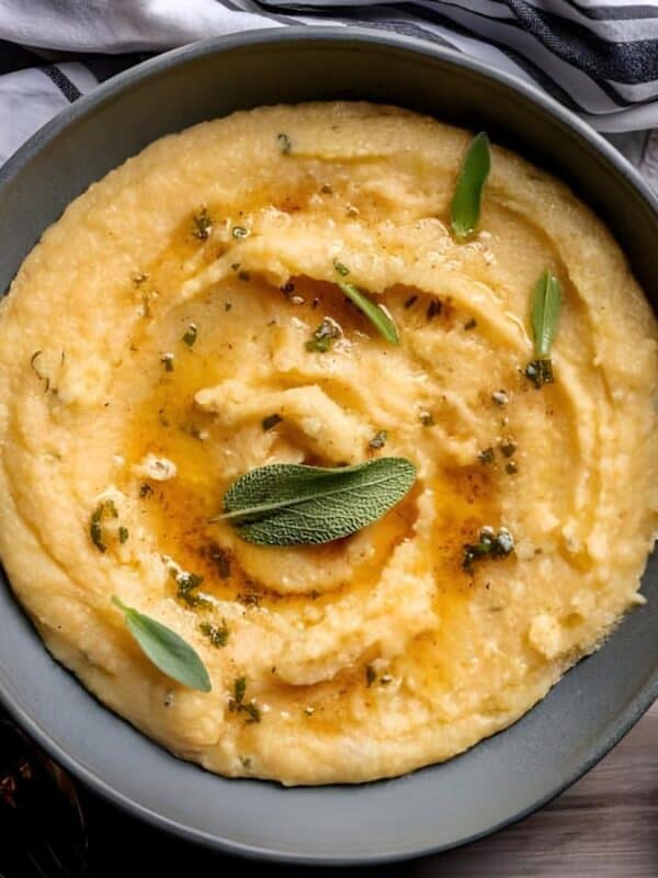 A bowl of creamy polenta topped with melted butter and fresh sage leaves, placed on a wooden tray with a striped napkin, gold utensils, and a small bowl of grated cheese on the side.