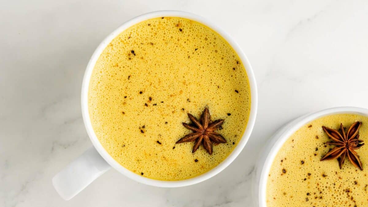 Two white mugs filled with frothy golden milk, each topped with a star anise, sit on a white marble surface.