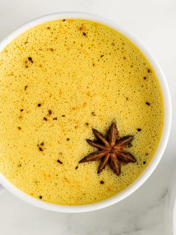 Two white mugs filled with frothy golden milk, each topped with a star anise, sit on a white marble surface.