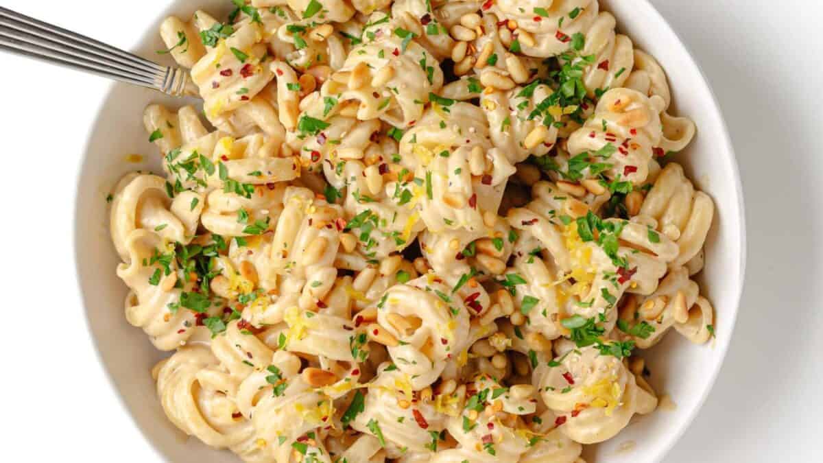 A bowl of creamy pasta topped with chopped parsley, red pepper flakes, pine nuts, and lemon zest, with a fork resting in the dish.