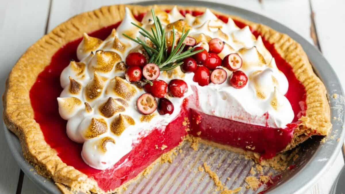 A cranberry pie topped with toasted meringue and garnished with fresh cranberries and rosemary. A large slice has been removed, revealing the bright red filling inside the pie crust.