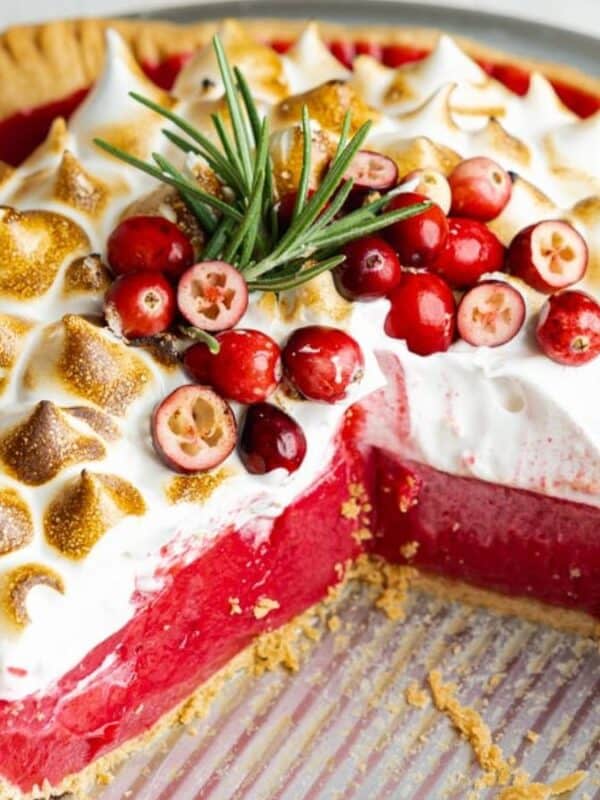 A cranberry pie topped with toasted meringue and garnished with fresh cranberries and rosemary. A large slice has been removed, revealing the bright red filling inside the pie crust.