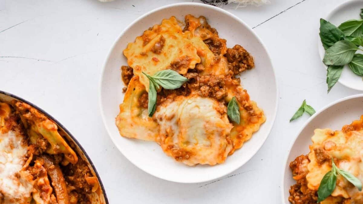 A white plate with cheesy baked ravioli in meat sauce, garnished with fresh basil leaves, placed on a white surface near a pot of more ravioli and a small plate of basil.