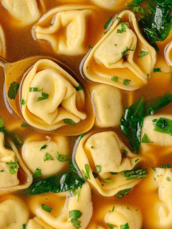 A bowl of tortellini soup with spinach, topped with chopped parsley. A spoon is lifting several tortellini from the clear, golden broth.