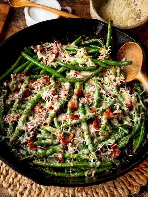 A skillet filled with green beans, bacon, and grated cheese sits on a woven placemat, surrounded by fresh green beans, a wedge of cheese, a grater, an onion, garlic, and a wooden spoon.