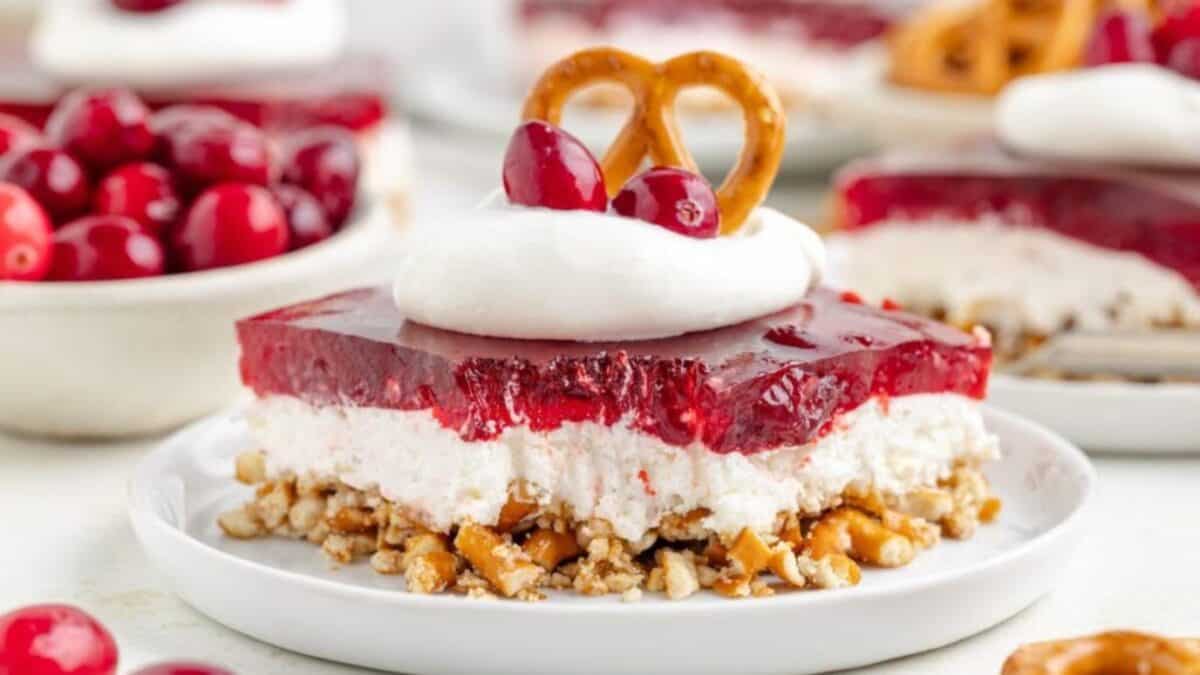 A dessert bar with a pretzel crust, creamy white layer, and red gelatin topping, garnished with whipped cream, whole cranberries, and a pretzel, served on a white plate. A bowl of cranberries is in the background.