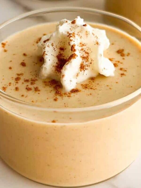 A glass bowl filled with creamy pumpkin mousse, topped with a dollop of whipped cream and a sprinkle of cinnamon, sits on a white marble surface.