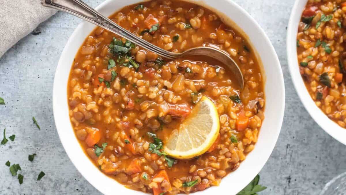 A bowl of lentil soup with vegetables, garnished with chopped herbs and a slice of lemon, with a spoon resting inside the bowl.