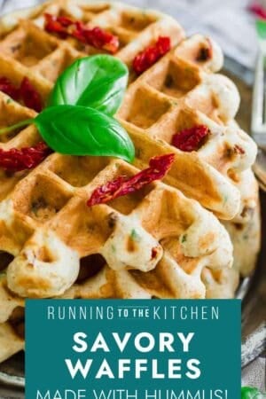 A stack of golden brown savory waffles garnished with fresh basil leaves and sun-dried tomatoes, with text overlay: "Running to the Kitchen. Savory Waffles Made with Hummus!.