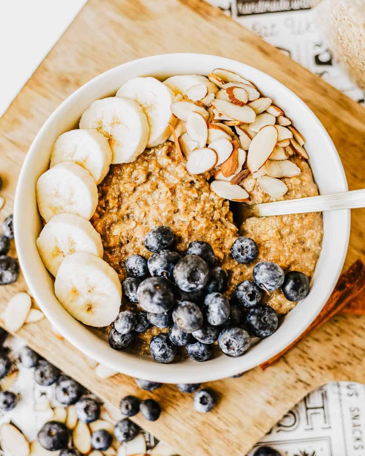 A bowl of instant brown sugar oatmeal topped with sliced bananas, fresh blueberries, and sliced almonds, placed on a wooden board with extra blueberries and almond slices scattered around.