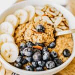 A bowl of brown sugar oatmeal topped with sliced bananas, fresh blueberries, sliced almonds, and a spoon resting inside the bowl.