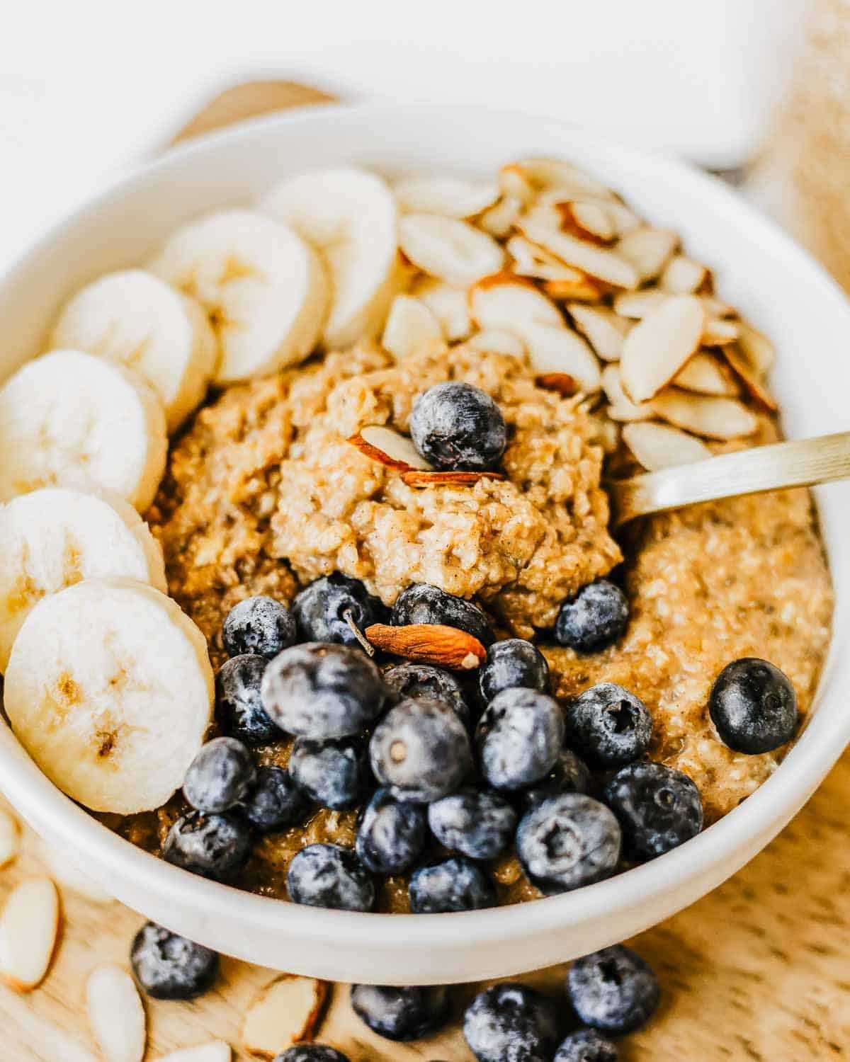 A bowl of brown sugar oatmeal topped with sliced bananas, fresh blueberries, sliced almonds, and a spoon resting inside the bowl.
