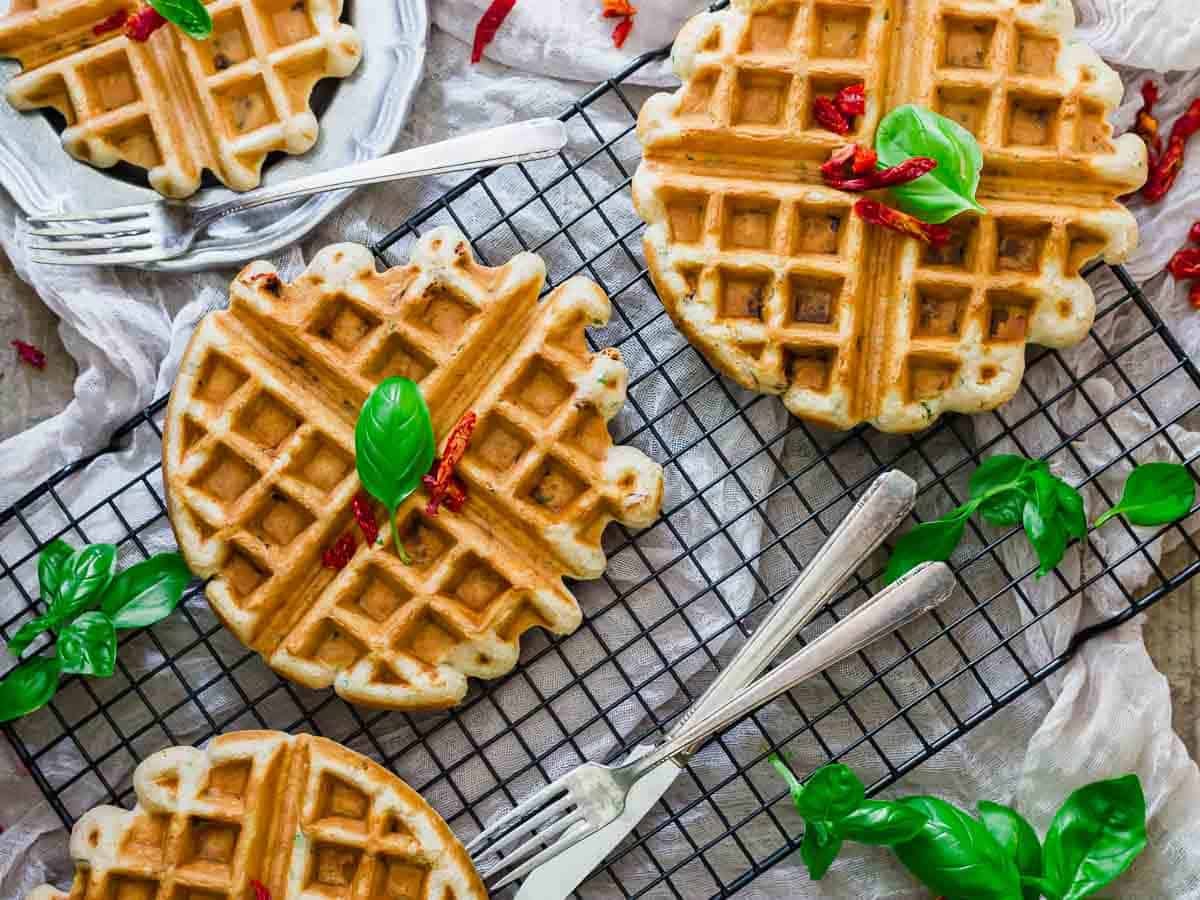 Four golden brown waffles on a cooling rack, garnished with fresh basil leaves and small red chili slices. Two forks are placed next to the waffles, and extra basil leaves are scattered around.