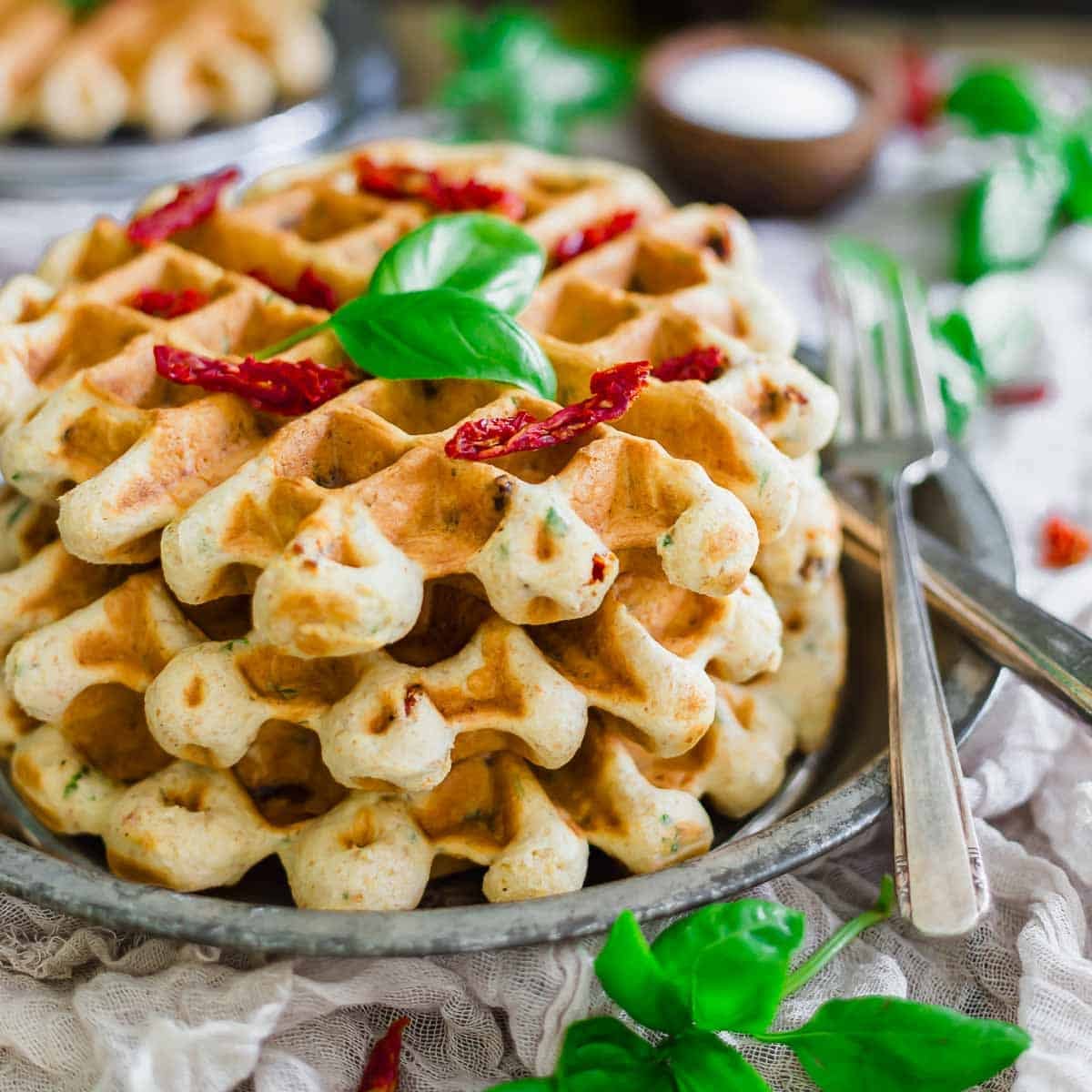 A stack of savory waffles garnished with fresh basil leaves and sun-dried tomatoes, served on a metal plate with a fork and knife, surrounded by scattered basil on a rustic table setting.
