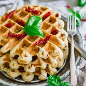 A stack of savory waffles garnished with fresh basil leaves and sun-dried tomato slices, served on a metal plate with a fork and knife, set on a rustic table with additional basil leaves scattered around.