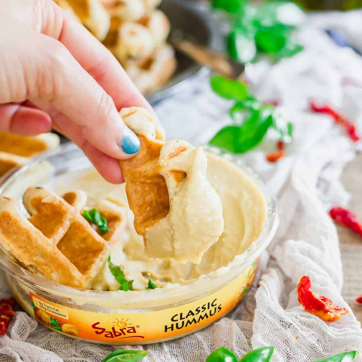 A hand with blue nail polish dips a piece of waffle into a container of Sabra Classic Hummus. Nearby, there are fresh herbs and sun-dried tomatoes on a light-colored surface.