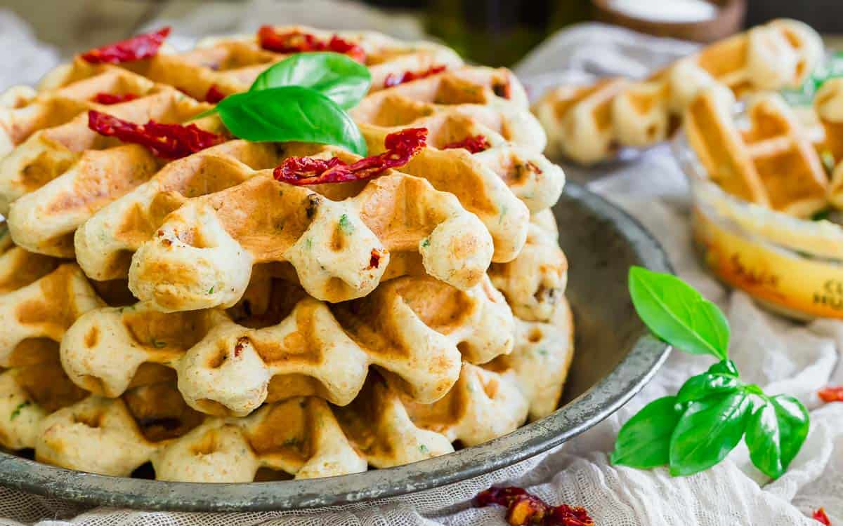 A stack of savory waffles garnished with sun-dried tomato slices and fresh basil leaves sits on a rustic plate, with more basil and waffle pieces in the background.