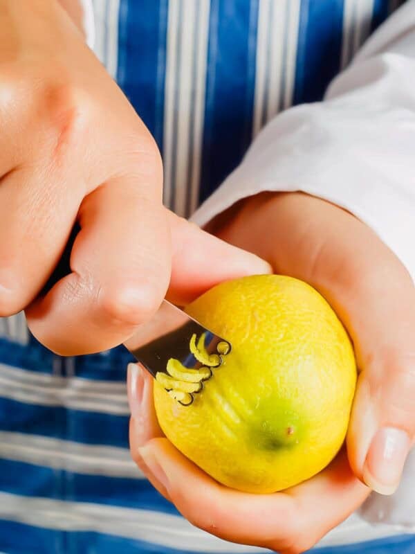 A person uses a small metal zester tool to remove thin strips of zest from a yellow lemon, with both hands visible. The person is wearing a white shirt and a blue striped apron.