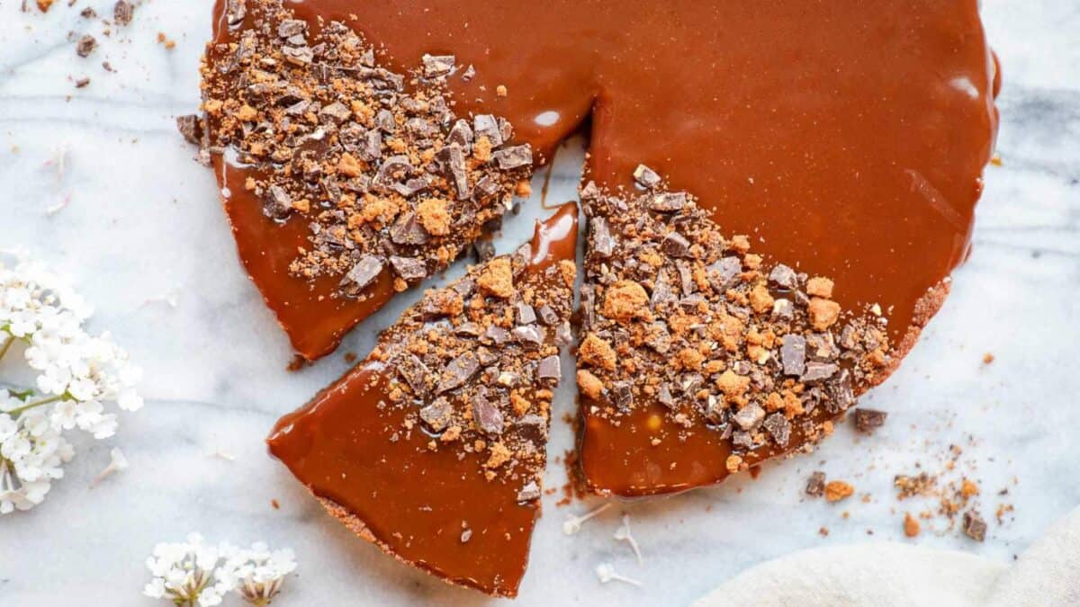 A chocolate tart with a smooth, glossy caramel topping, partially sliced. Some slices are topped with crushed chocolate pieces. The tart sits on a marble surface, with white flowers nearby.