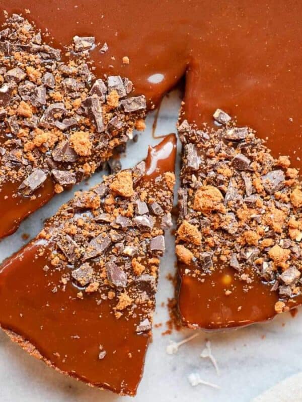 A chocolate tart with a smooth, glossy caramel topping, partially sliced. Some slices are topped with crushed chocolate pieces. The tart sits on a marble surface, with white flowers nearby.