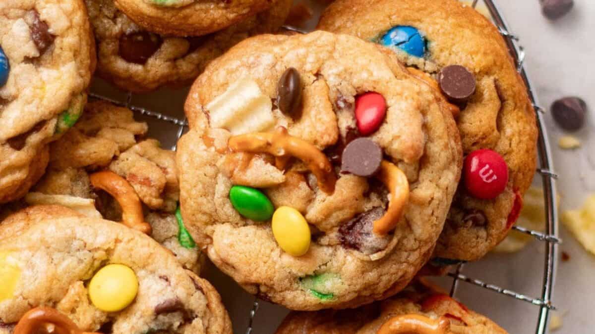A close-up of cookies topped with colorful chocolate candies, chocolate chips, broken pretzels, and bits of potato chips, resting on a round cooling rack.
