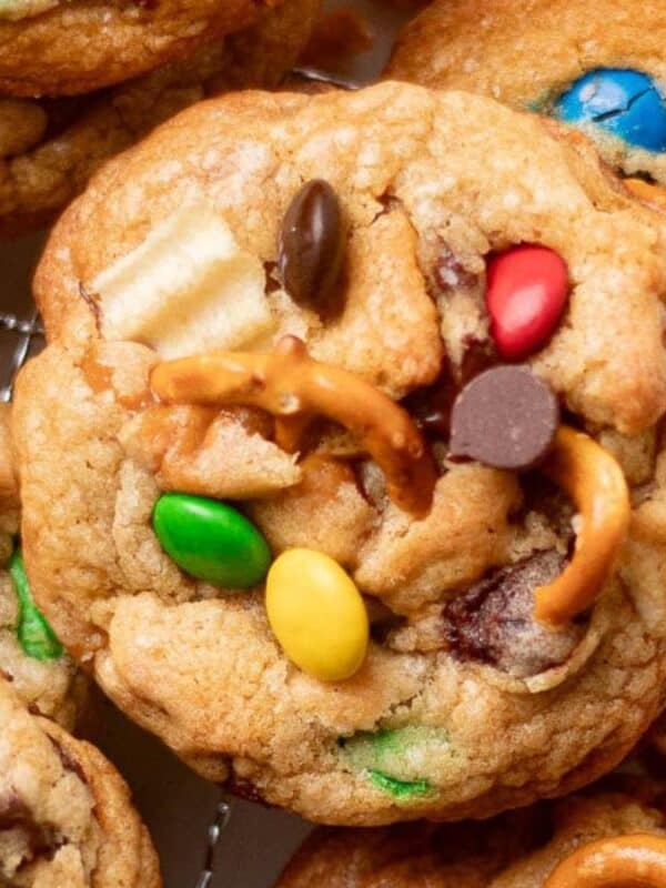 A close-up of cookies topped with colorful chocolate candies, chocolate chips, broken pretzels, and bits of potato chips, resting on a round cooling rack.