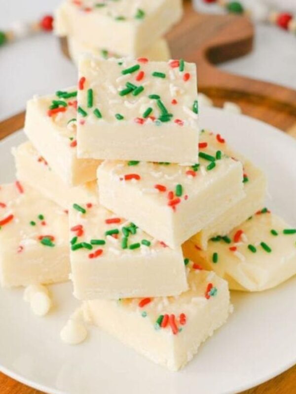 A plate of white chocolate fudge squares topped with red and green sprinkles is stacked on a wooden tray. Festive decorations and a holiday-themed cloth are in the background.