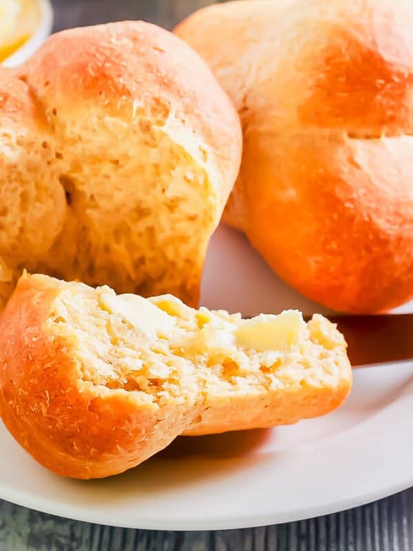A plate with two cloverleaf dinner rolls, one partially split open and spread with butter using a knife. A small bowl of butter is in the background on a wooden surface.