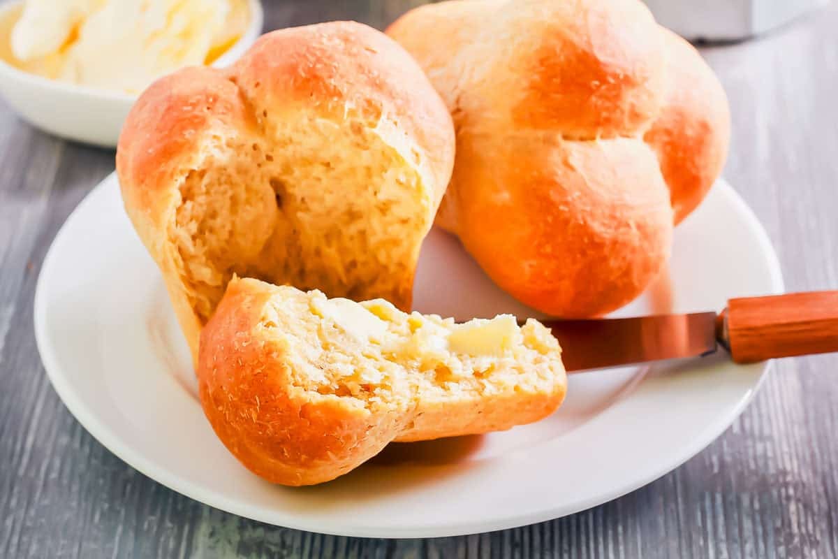 A plate with two cloverleaf dinner rolls, one partially split open and spread with butter using a knife. A small bowl of butter is in the background on a wooden surface.