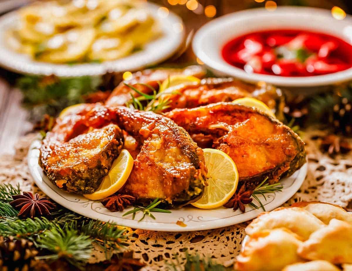 A plate of fried fish garnished with lemon slices and herbs, surrounded by festive decorations, with bowls of red soup and dumplings in the background.