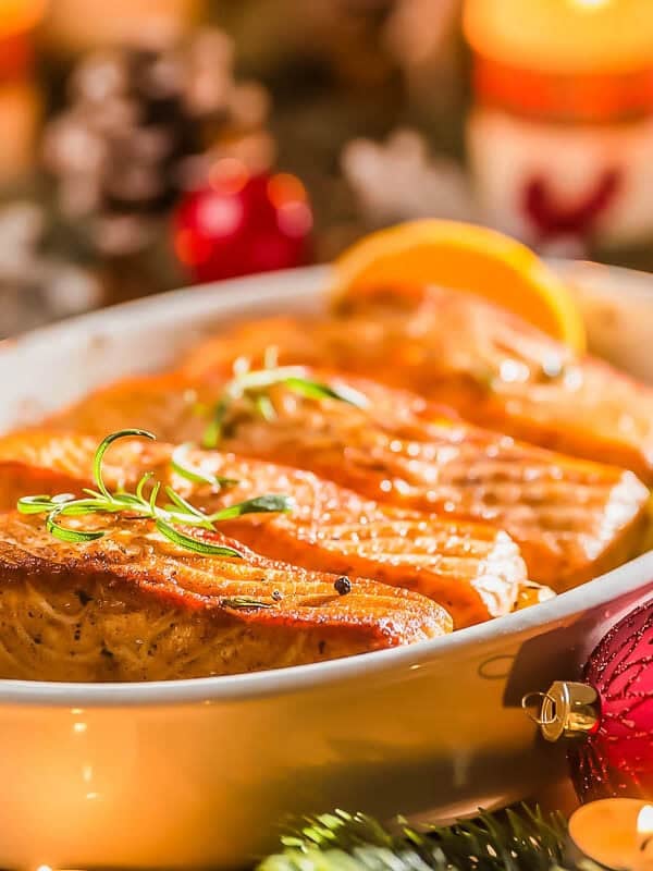 A festive dish of baked salmon fillets garnished with herbs is served in a white ceramic dish, surrounded by Christmas decorations, candles, pinecones, and ornaments.